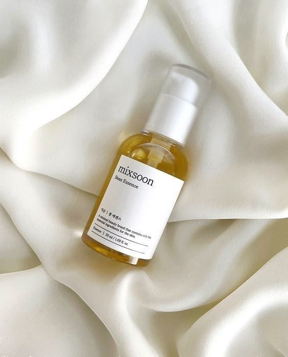 Saerún Skin: Botella de esencia Mixsoon Bean Essence con líquido dorado sobre tela blanca sedosa.